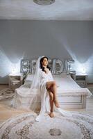 un incroyablement magnifique la mariée dans le Matin dans une blanc peignoir et une longue voile est assis sur le lit dans sa chambre à coucher. le la mariée pose dans le Matin avant le mariage cérémonie. photo