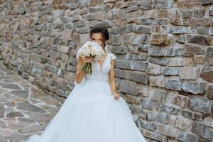 portrait. une brunette la mariée dans une robe et une voile, posant avec une bouquet contre le Contexte de une pierre mur. argent bijoux. magnifique maquillage et cheveux. l'automne mariage. fête photo