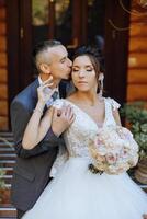 une Jeune la mariée et jeune marié tendrement embrasse dans le des rayons de le l'automne Soleil. soumissionner et magnifique Jeune fille la mariée. une homme baisers le sien bien-aimé. contre le Contexte de une magnifique jardin photo