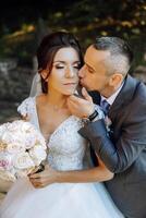 une Jeune la mariée et jeune marié tendrement embrasse dans le des rayons de le l'automne Soleil. soumissionner et magnifique Jeune fille la mariée. une homme baisers le sien bien-aimé. contre le Contexte de une magnifique jardin photo