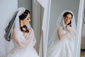 une Jeune européen femme avec foncé cheveux dans une mariage robe des stands par une miroir dans une studio décoré avec fleurs. Jeune magnifique la mariée. photo