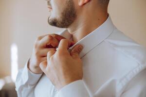 une homme dans une blanc chemise des stands par le fenêtre dans le pièce et attache le boutons sur le sien collier et manches. regarder sur main. élégant affaires portrait de une homme, fermer photo. le jeune marié est en train de préparer. photo