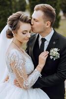 une Jeune la mariée et jeune marié tendrement embrasse dans le des rayons de le l'automne Soleil. soumissionner et magnifique Jeune fille la mariée. une homme baisers le sien bien-aimé. contre le Contexte de une magnifique jardin photo