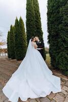 une Jeune la mariée et jeune marié tendrement embrasse dans le des rayons de le l'automne Soleil. soumissionner et magnifique Jeune fille la mariée. une homme baisers le sien bien-aimé. contre le Contexte de une magnifique jardin photo