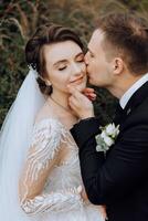 une Jeune la mariée et jeune marié tendrement embrasse dans le des rayons de le l'automne Soleil. soumissionner et magnifique Jeune fille la mariée. une homme baisers le sien bien-aimé. contre le Contexte de une magnifique jardin photo