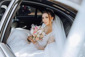 le la mariée est séance dans une noir voiture avec une bouquet de fleurs dans sa mains et regards en dehors de le auto. portrait de une plutôt timide la mariée dans une auto. mariée, sourire, émotions. photo