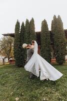 Beau jeune marié porter une magnifique en riant la mariée dans le sien bras dans une printemps parc. le jeune marié est encerclant avec le la mariée dans le sien bras photo