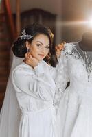 portrait de un incroyablement magnifique fille la mariée dans une blanc peignoir dans le chambre à coucher, le la mariée pose dans le Matin avant le mariage cérémonie. photo