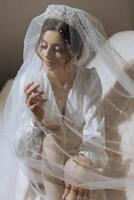 portrait de un incroyablement magnifique fille la mariée dans une blanc peignoir dans le chambre à coucher, le la mariée pose en portant sa voile dans sa mains et couvertures se avec il. photo