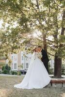 mariage portrait. une jeune marié dans une noir costume et une blond la mariée sont debout, étreindre et embrasser en dessous de une arbre. photo session dans la nature. une magnifique rayon de le Soleil dans le photo. magnifique cheveux et maquillage