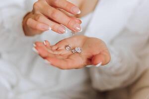 le la mariée détient dans sa mains et spectacles sa des boucles d'oreilles avec précieux des pierres, fermer. Matin de le la mariée. le mariage journée. photo