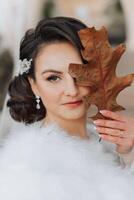 mariage portrait. une brunette la mariée avec un ornement dans sa cheveux pose en portant une feuille près sa affronter. l'automne mariage. magnifique maquillage et cheveux. préparation pour le fête photo