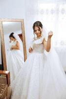 une Jeune européen femme avec foncé cheveux dans une mariage robe des stands par une miroir dans une studio décoré avec fleurs. Jeune magnifique la mariée. photo