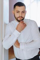 une homme dans une blanc chemise des stands par le fenêtre dans le pièce et attache le boutons sur le sien collier et manches. regarder sur main. élégant affaires portrait de une homme, fermer photo. le jeune marié est en train de préparer. photo