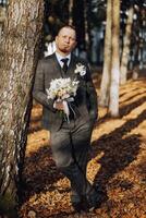 portrait de une Beau Jeune jeune marié sur un l'automne journée en plein air dans un élégant costume et en portant une mariage bouquet de fleurs. Extérieur photo. Beau homme dans une affaires costume. photo