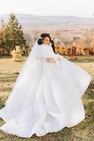 une brunette la mariée dans une blanc robe avec une longue train détient le robe et des promenades vers le bas le chemin couvert avec l'automne feuilles. mariage photo session dans la nature. magnifique cheveux et se maquiller. fête