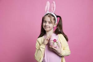 joyeux Jeune enfant portant duveteux lapin oreilles pose avec rose œuf, en présentant sa Fait main peint Pâques ornement plus de rose Contexte. content petit fille célébrer avril vacances festivité. photo