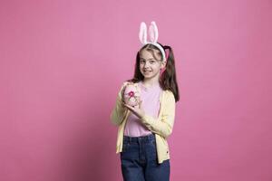 petit charmant enfant avec lapin oreilles en portant une peint rose Oeuf dans de face de caméra, en présentant sa Fait main Pâques décoration plus de coloré Contexte. Jeune adorable fille spectacles traditionnel ornements. photo