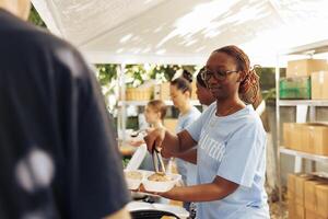 africain américain femme portion gratuit nourriture à le pauvre, nécessiteux individuel à un Extérieur nourriture banque. femelle bénévoles alimentation et fournir soutien à le faim et Moins privilégié à une sans abri abri. photo