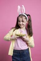 charmant peu enfant avec lapin oreilles pose avec adorable printemps jouets tandis que porter un Pâques panier plein de des œufs et une poussin. joyeux et enthousiaste enfant profiter le vacances dans studio. photo