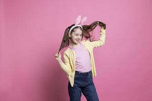 souriant sur de soi bambin dansant autour dans le studio avec lapin oreilles, sentiment content et excité à propos Pâques un événement vacances. Jeune enfant étant joyeux contre rose arrière-plan, portant nattes. photo