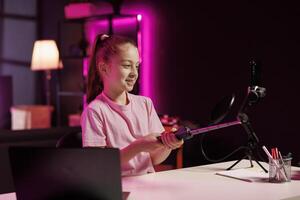 mignonne fille tournage pièce tour à l'intérieur Accueil studio en utilisant téléphone intelligent sur selfie bâton. peu enfant Faire vidéo en présentant sa néon allumé appartement à intéressé les enfants public photo