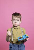 mécontent irrité petit garçon sentiment triste tandis que il pièces avec une jouet voiture, presque pleurs dans de face de le caméra. peu enfant sentiment en colère et dérangé en portant une Plastique bleu voiture. photo