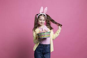 dans le studio, une enfant exprime sa enthousiasme pour Pâques par en portant coloré jouets pour le caméra. peu adorable enfant en portant une farci lapin et un des œufs panier, portant lapin oreilles. photo