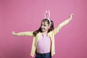 joyeux petit enfant avec nattes dansant autour dans de face de caméra, montrer du doigt à quelque chose dans studio et ayant amusement. Jeune bambin sentiment sur de soi tandis que elle est portant lapin oreilles. photo