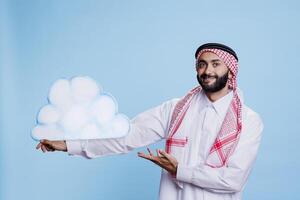 souriant musulman homme en portant nuage forme et montrer du doigt avec main studio portrait. la personne habillé dans arabe thobe et foulard traditionnel vêtements en présentant temps prévoir et à la recherche à caméra photo