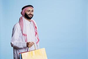 souriant musulman homme en portant Jaune achats sac et à la recherche à caméra avec de bonne humeur expression. arabe Shopaholic portant traditionnel vêtements porter achat studio portrait photo