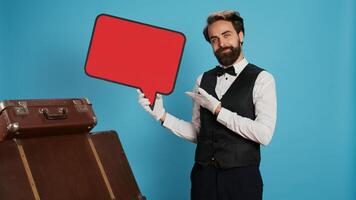 Hôtel porter en portant discours bulle sur publicité, en présentant Vide fond sur panneau d'affichage. élégant chic portier mettant en valeur vide bannière papier carton signe sur caméra contre bleu Contexte. photo