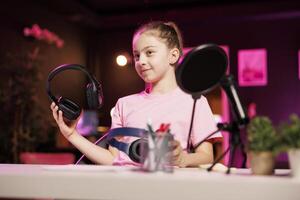 enfant présente écouteurs dans différent prix gammes à public, comparant leur à trouver gagnant, tournage dans néon allumé Accueil studio. gen z technologie influenceur enfant recommande écoute dispositifs à les spectateurs photo