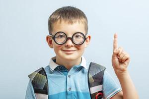 une mignonne école garçon dans des lunettes avec élevé indice doigt sur bleu Contexte photo