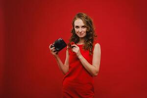 une souriant Jeune femme avec ondulé cheveux détient une fraise et photographies il, en portant une délicieux Frais fraise sur une brillant rouge Contexte photo