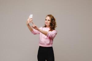 une souriant content fille dans une rose chemisier prend une selfie sur une téléphone intelligent sur une gris Contexte. photo