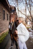 Jeune cavalier femme blond avec longue cheveux dans blanc vêtements permanent près marron cheval photo