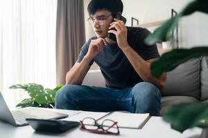 le homme dans décontractée vêtements travail avec une ordinateur portable, ordinateur, intelligent téléphone, calculatrice séance sur le canapé dans le vivant pièce à maison, travail de Accueil concept. photo