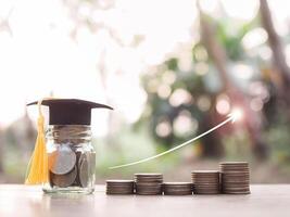 verre bouteille avec l'obtention du diplôme chapeau et empiler de pièces de monnaie avec La Flèche en hausse Icônes. le concept de économie argent pour éducation, étudiant prêt, bourse, frais de scolarité honoraires photo