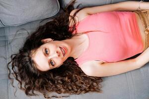 Jeune femme avec longue marron cheveux mensonges avec une sourire sur le canapé dans une rose T-shirt et dans croisillons, Haut vue photo