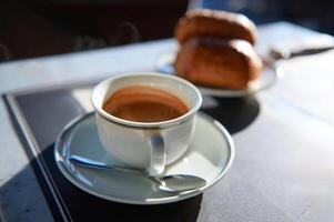 élégant blanc céramique tasse de fraîchement brassée Expresso café avec mousse sur soucoupe. flou croustillant des croissants sur Contexte photo