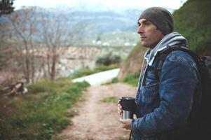 détendu content voyageur promeneur homme en portant une Voyage thermos agresser et à la recherche loin, permanent sur le forêt chemin dans montagnes photo