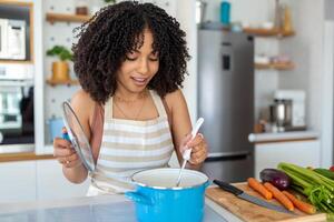 photo de jolie africain américain femme en portant cuisine louche cuillère tandis que cuisine soupe avec Frais des légumes dans cuisine à Accueil