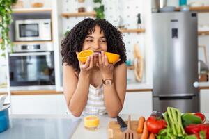 photo de mignonne africain américain femme souriant et en portant deux Orange les pièces tandis que cuisine légume salade dans cuisine intérieur à Accueil