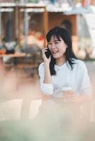 une Jeune femme actions une joyeux conversation sur sa mobile téléphone, sa sourire rayonnant bonheur, tandis que en portant une à emporter café tasse en plein air, illustrant une détendu mode de vie. photo