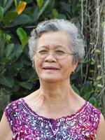 une Sénior asiatique femme avec court blanc cheveux souriant et à la recherche à le caméra tandis que permanent dans une jardin photo