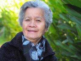 Sénior asiatique femme avec court blanc cheveux souriant et à la recherche à le caméra tandis que permanent dans une jardin. espace pour texte. concept de vieilli gens et soins de santé photo