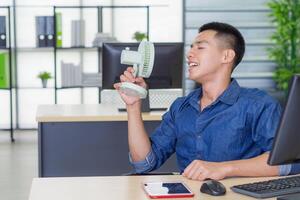 Jeune asiatique homme en portant une petit Plastique ventilateur soufflant à le sien visage car de chaud temps tandis que séance dans une chaise dans le bureau. espace pour texte. concept de gens et relaxation photo