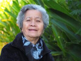 Sénior asiatique femme avec court blanc cheveux souriant et à la recherche à le caméra tandis que permanent dans une jardin. espace pour texte. concept de vieilli gens et soins de santé photo