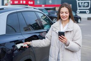 Jeune femme mise en charge sa électrique voiture à une mise en charge station dans le ville. éco carburant concept. le concept de écologiquement amical transport. recharger batterie de mise en charge gare. photo